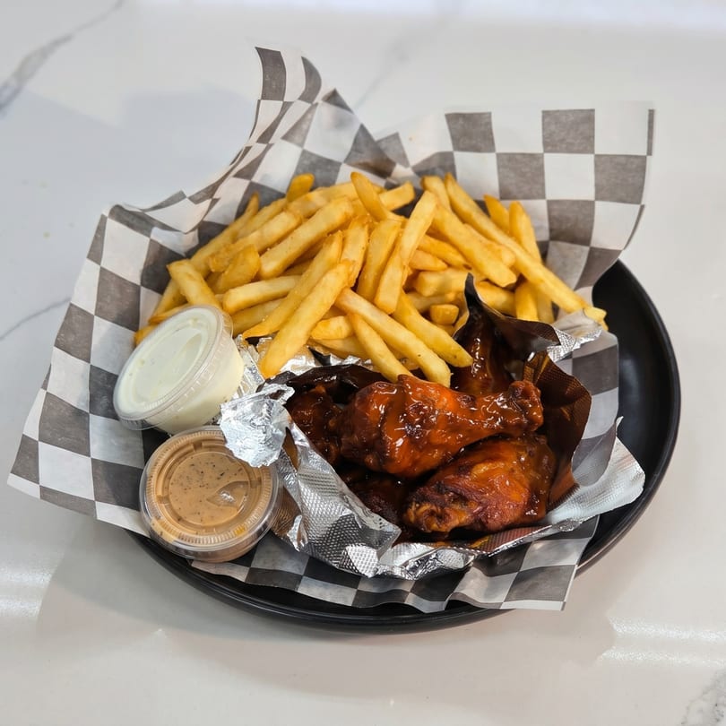 A black plate holds a basket of golden french fries, BBQ chicken wings, and two dipping sauces.