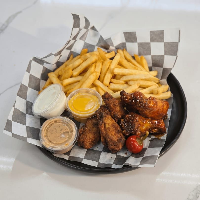A black tray with checkered paper holds french fries, chicken wings, and three dipping sauces.