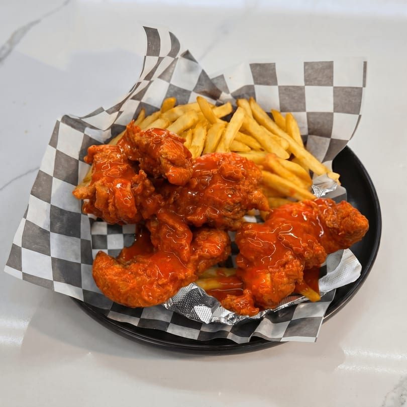 Crispy chicken tenders drenched in hot sauce, served with golden french fries on a black plate.