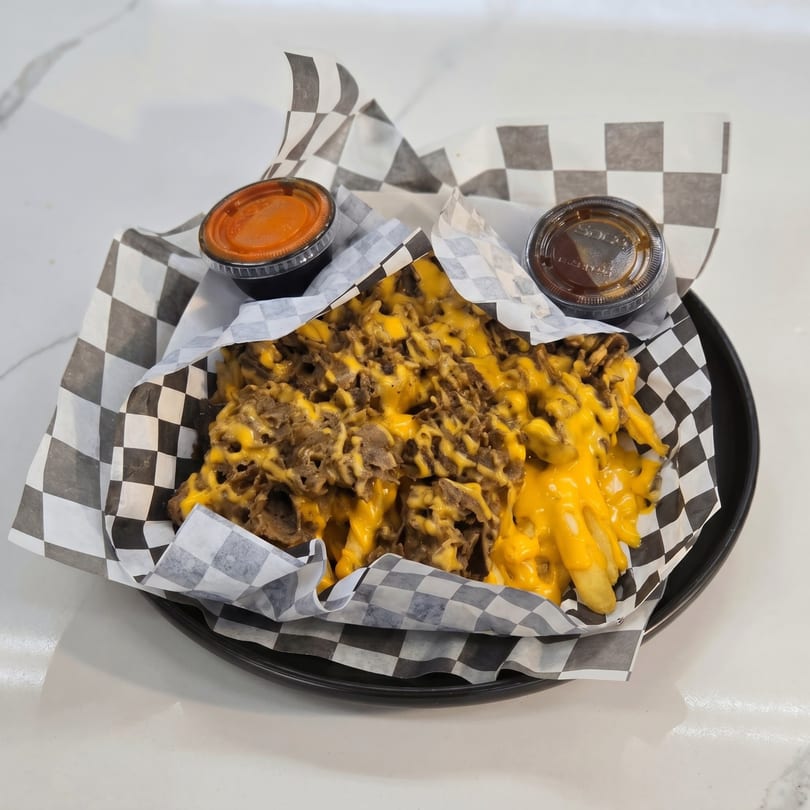 Cheesesteak fries with melted cheese and shredded steak, served with two dipping sauces in checkered paper.